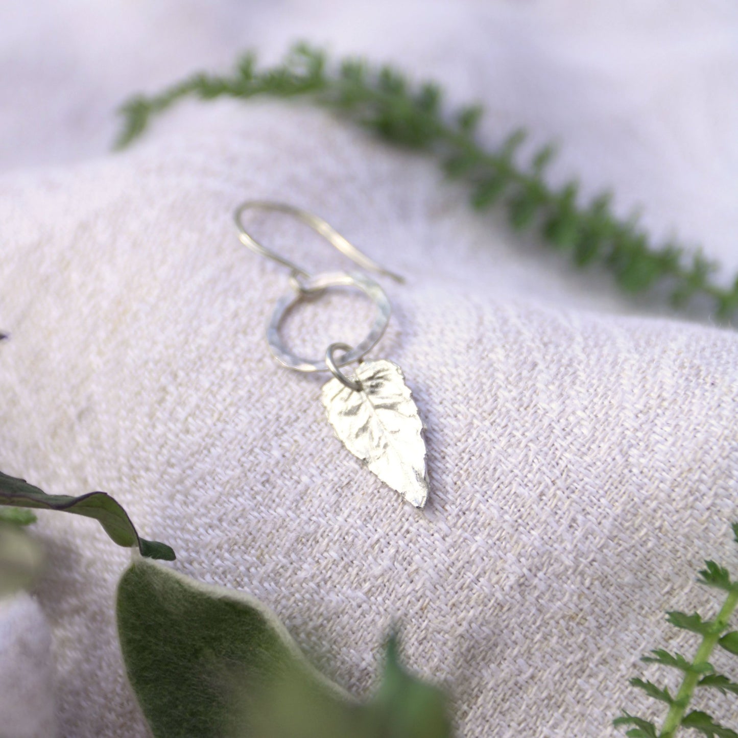 Silver leaf earring on a white fabric background with green leaves.