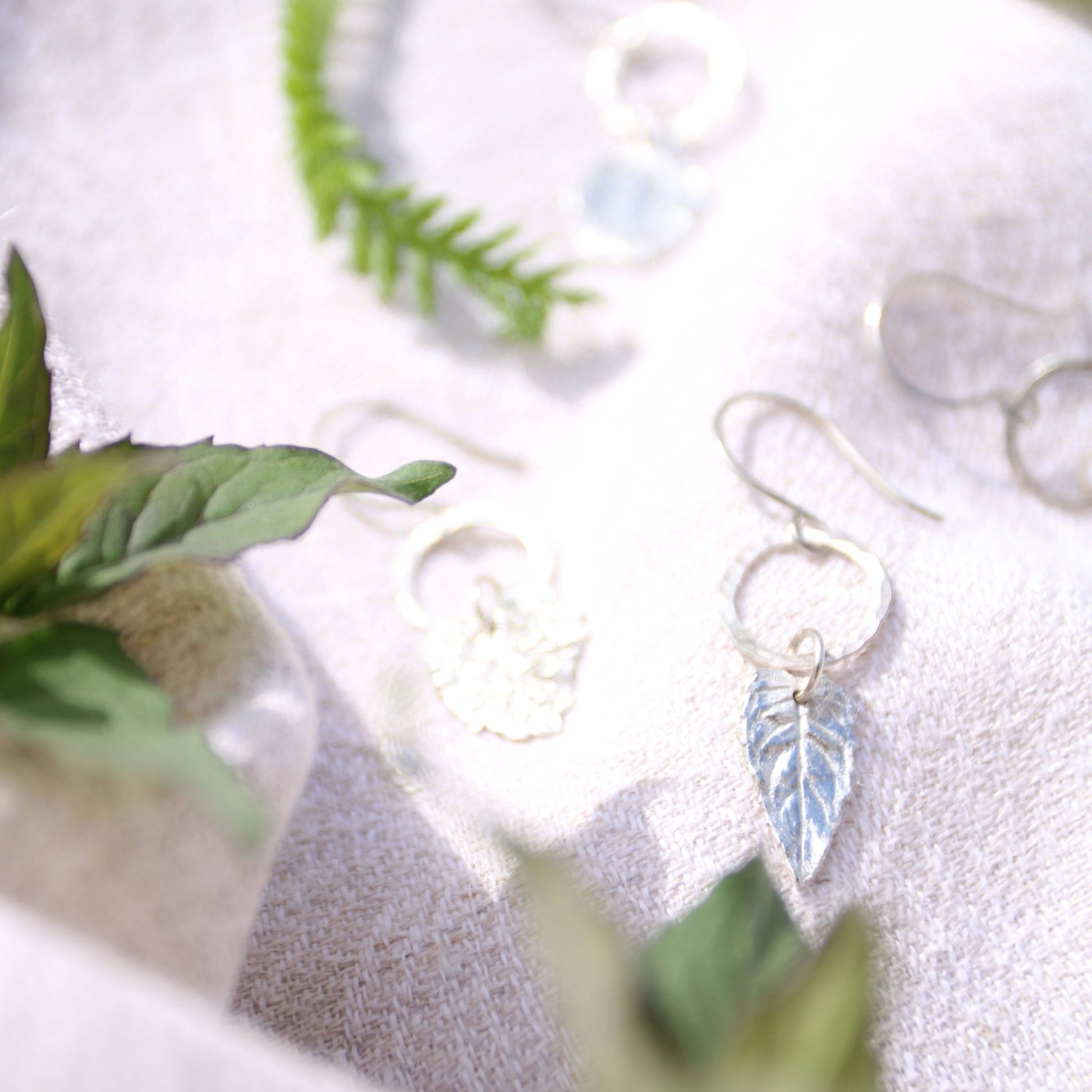 Silver earrings with leaf charms on a white surface with green leaves.
