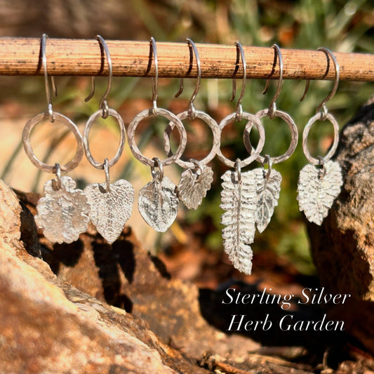 Silver herb garden clips with leaf designs hanging on a stick against a natural background.
